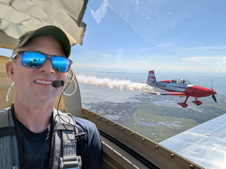 A pilot smiling in the cockpit while a red RV flies nearby trailing smoke.